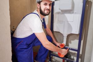 Handsome bearded man in work overalls installing concealed toilet frame in bathroom. Cheerful male worker in safety helmet looking at camera and smiling while installing wall-hung toilet system.